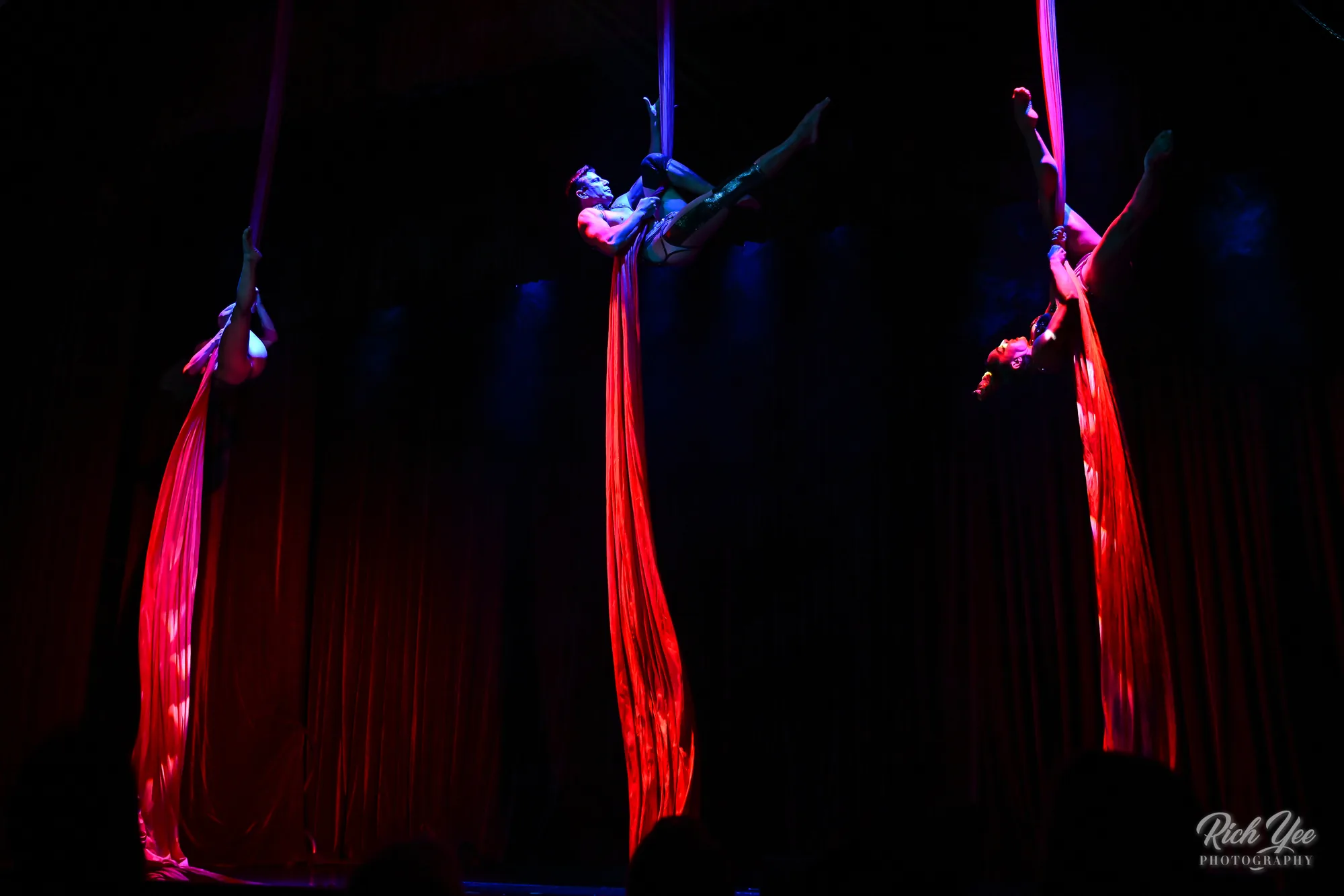 Three aerialists on red silks against a black stage